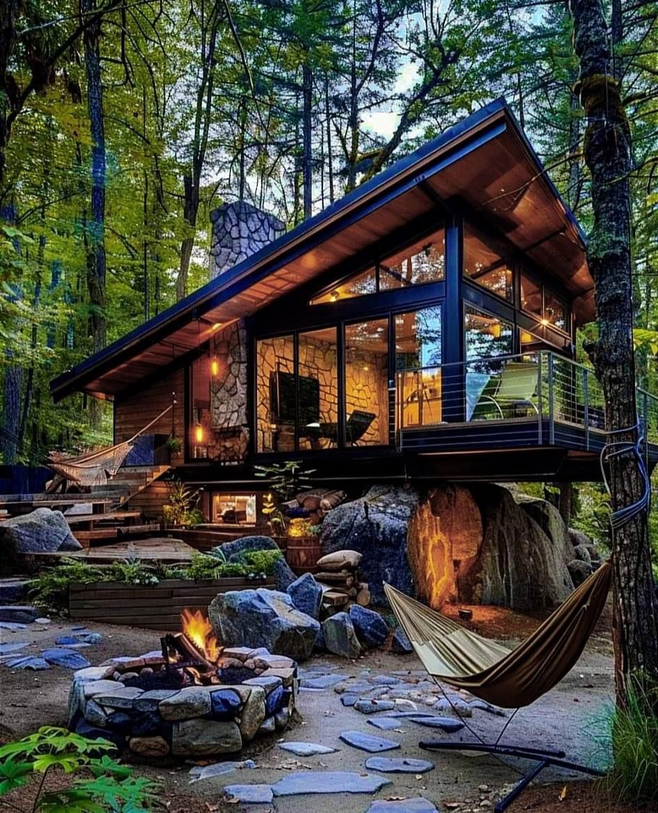 Glass-and-timber cabin in the forest with a stone firepit and hammock at dusk